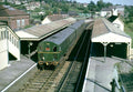 Green train on tracks at a station with buildings and trees in the background