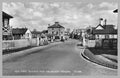 Vintage black and white photo of a town street with buildings and people from a railway crossing perspective.
