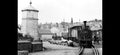 Vintage black and white photograph of a steam train on tracks with a small building and people in the background.