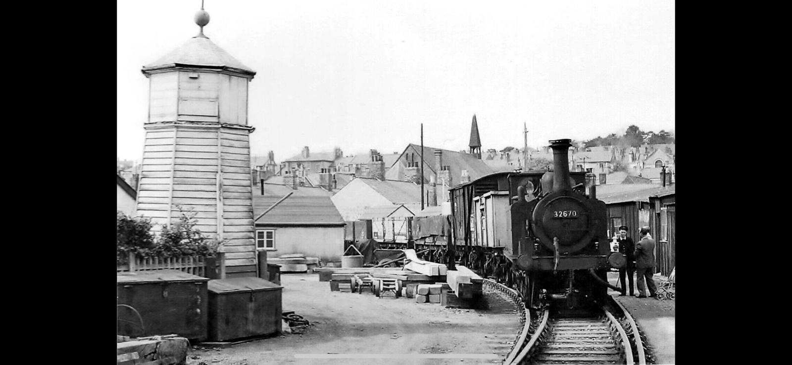 Vintage black and white photograph of a steam train on tracks with a small building and people in the background.