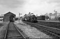 Vintage black and white photograph of a steam train at a station with a building and tracks in the foreground.