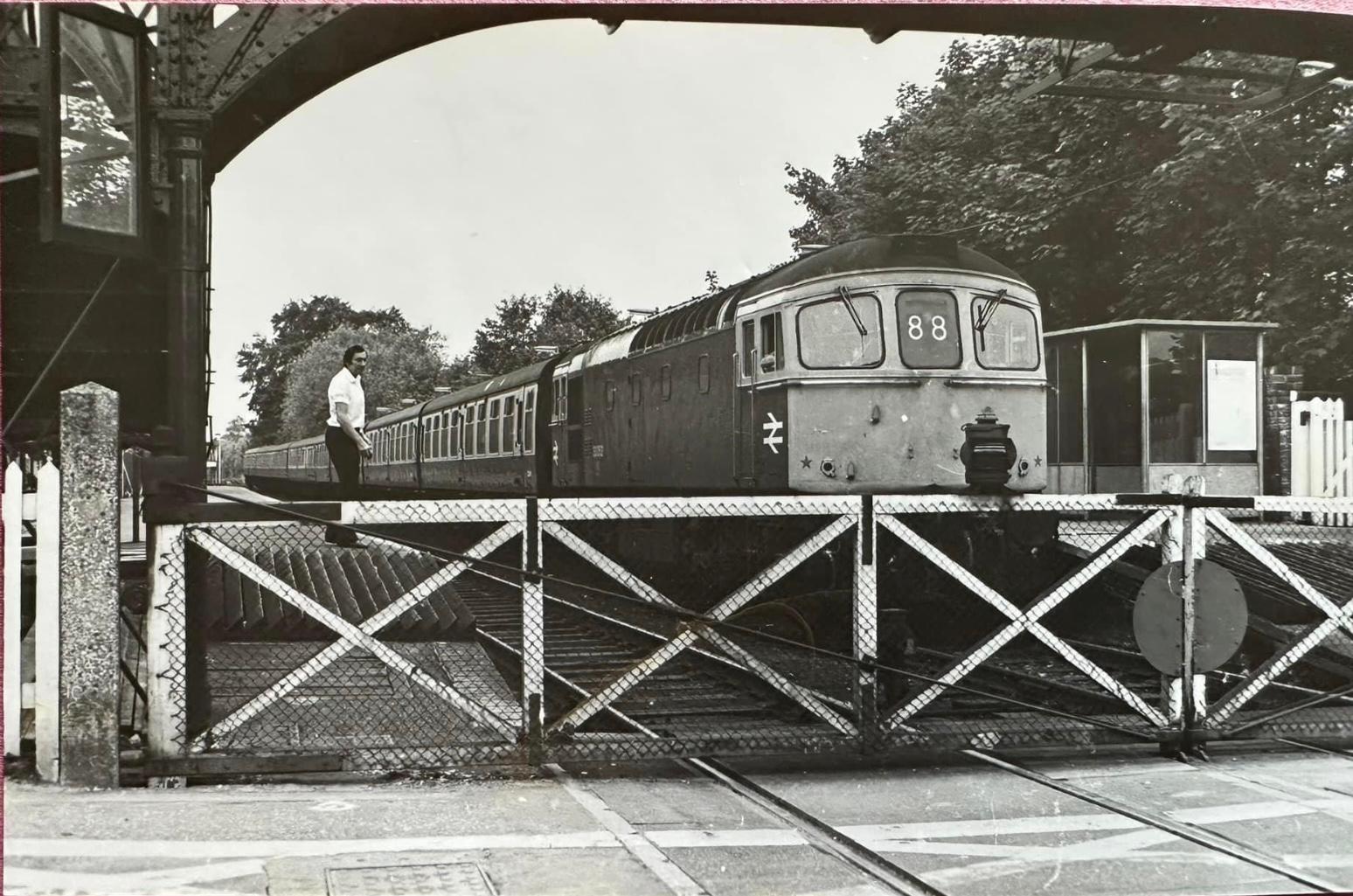 Vintage black and white photograph of a train crossing a level crossing with a person standing nearby.